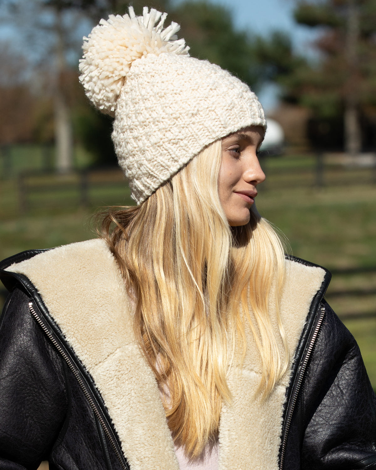 Woman wearing Eco Poppy Beanie in Arctic Wolf with pom-pom and a chunky knit design, standing outdoors in a black and beige jacket.